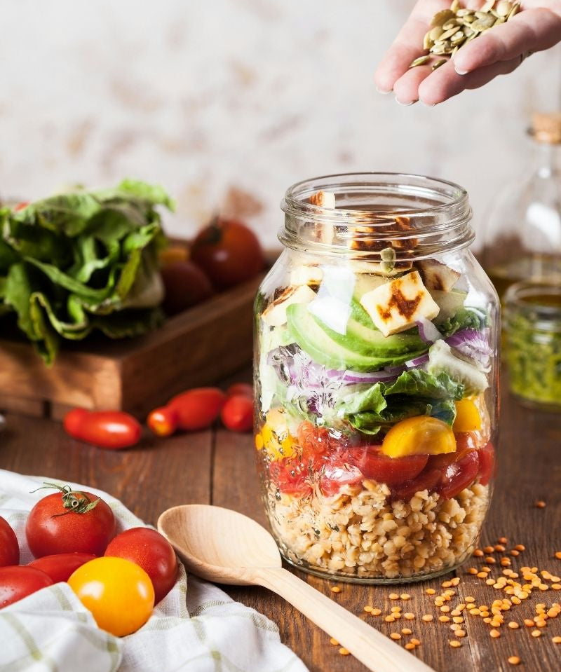 A big glass jar filled with a layered salad. Someone is sprinkling seeds into the jar.